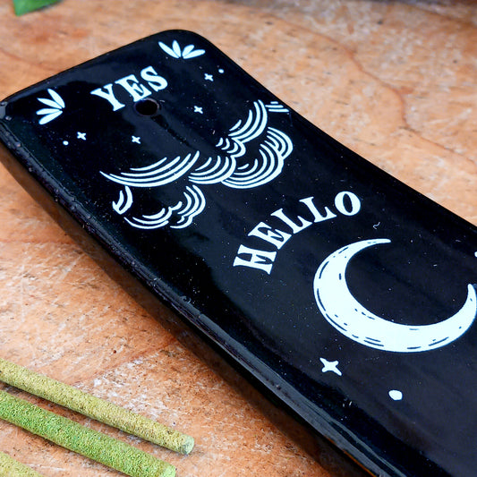 Add a touch of mystical gothic style to your incense rituals with this striking black incense ash catcher, inspired by traditional talking boards. Featuring a decorative planchette-style design surrounded by stars, moons and an all-seeing eye, it creates an atmospheric setting for burning your favourite incense sticks.