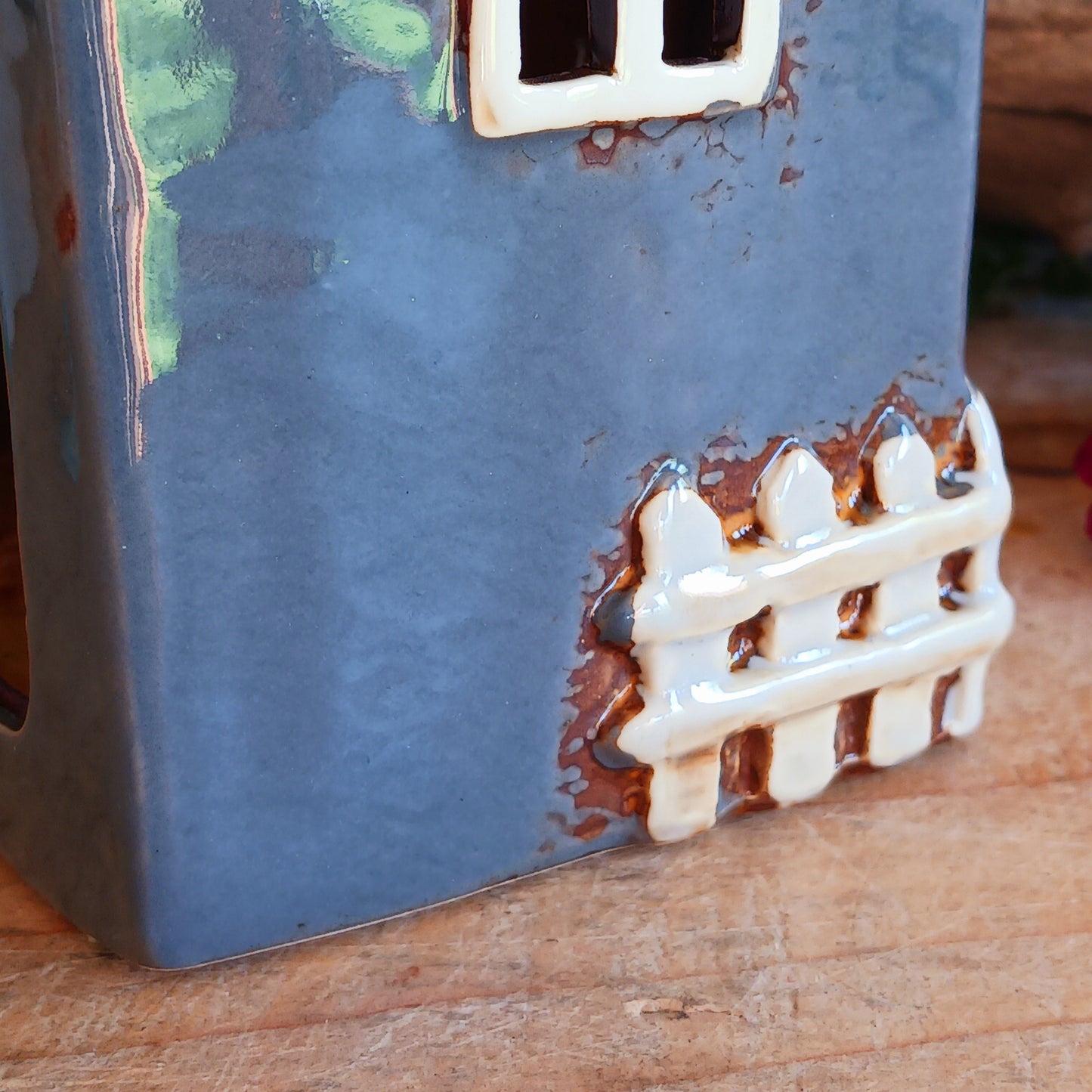 A beautiful ceramic garden house tealight holder crafted and hand-finished by Village Pottery making each one unique and when a tealight is lit it's cosy light is visible through the windows. This lovely ceramic house is decorated with a flowering garden pot plant and picket fence by the front door and coloured in navy blue with a cream tiled roof and red door.