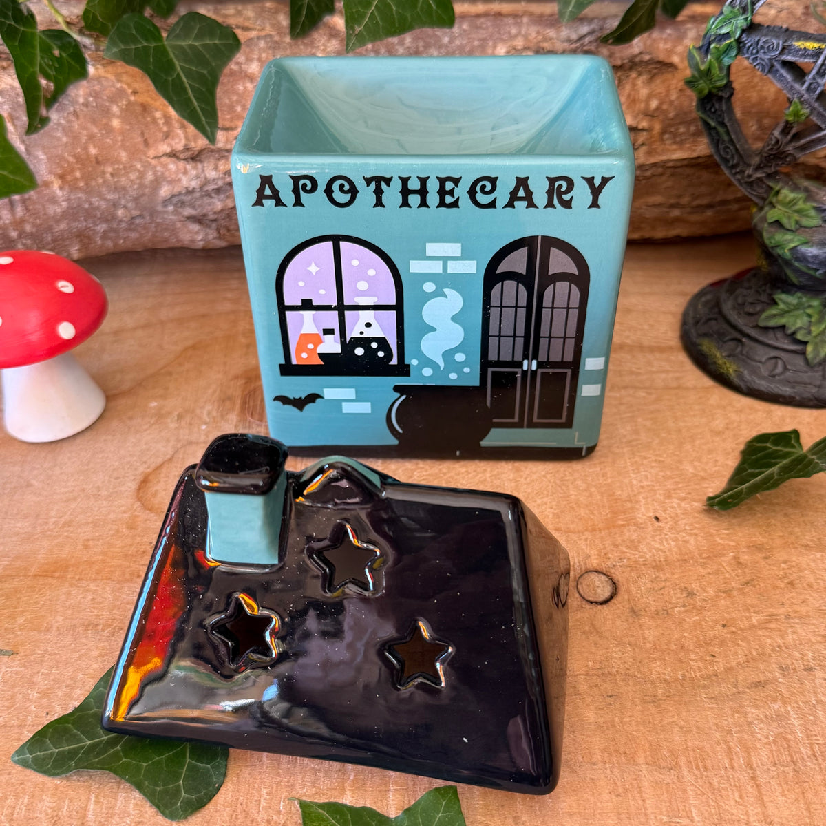 A Halloween-themed ceramic apothecary set on a wooden surface, featuring a light blue box printed with an apothecary shop design and a glossy black star-shaped lid with a small matching blue top piece.