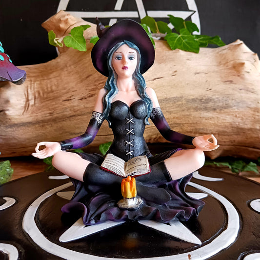 This stunning figurine features Aradia, wearing a black corseted dress, sitting crossed legged, cradling her grimoire with her legs, while staring into the burning flame of a candle.