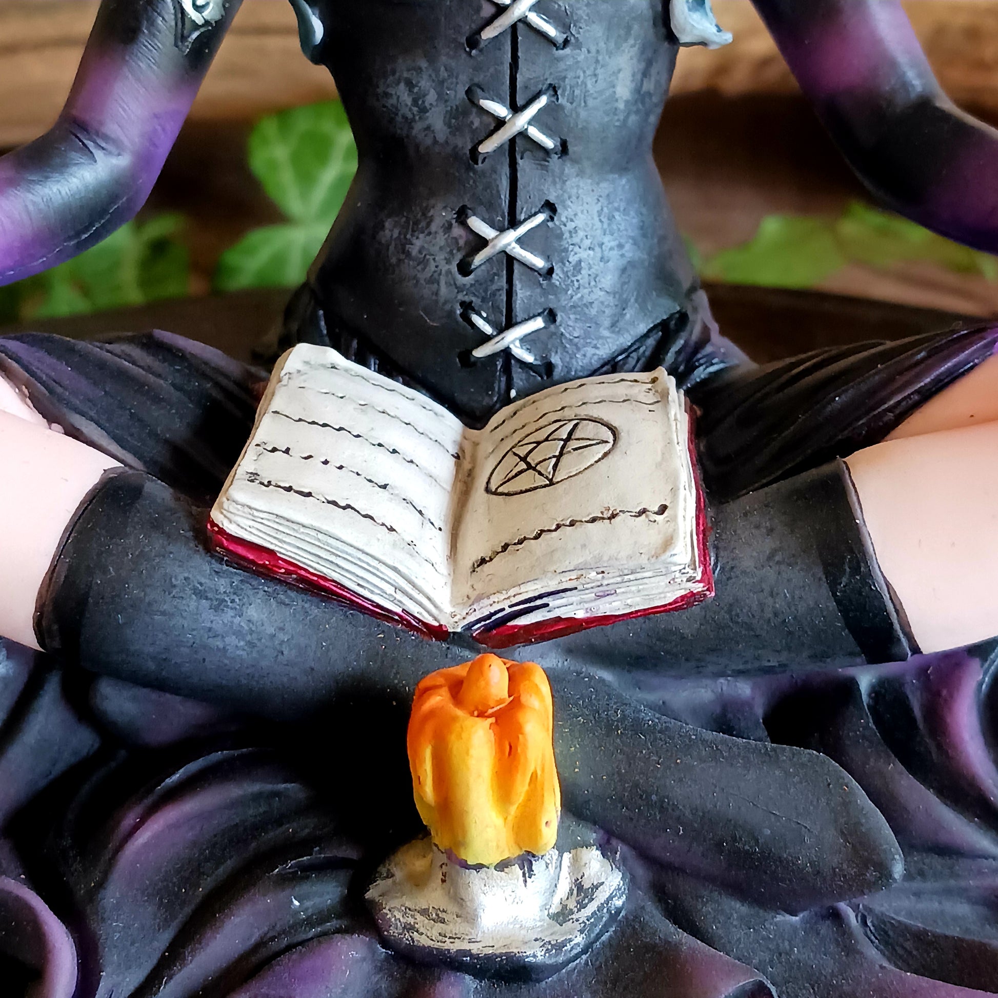 This stunning figurine features Aradia, wearing a black corseted dress, sitting crossed legged, cradling her grimoire with her legs, while staring into the burning flame of a candle.