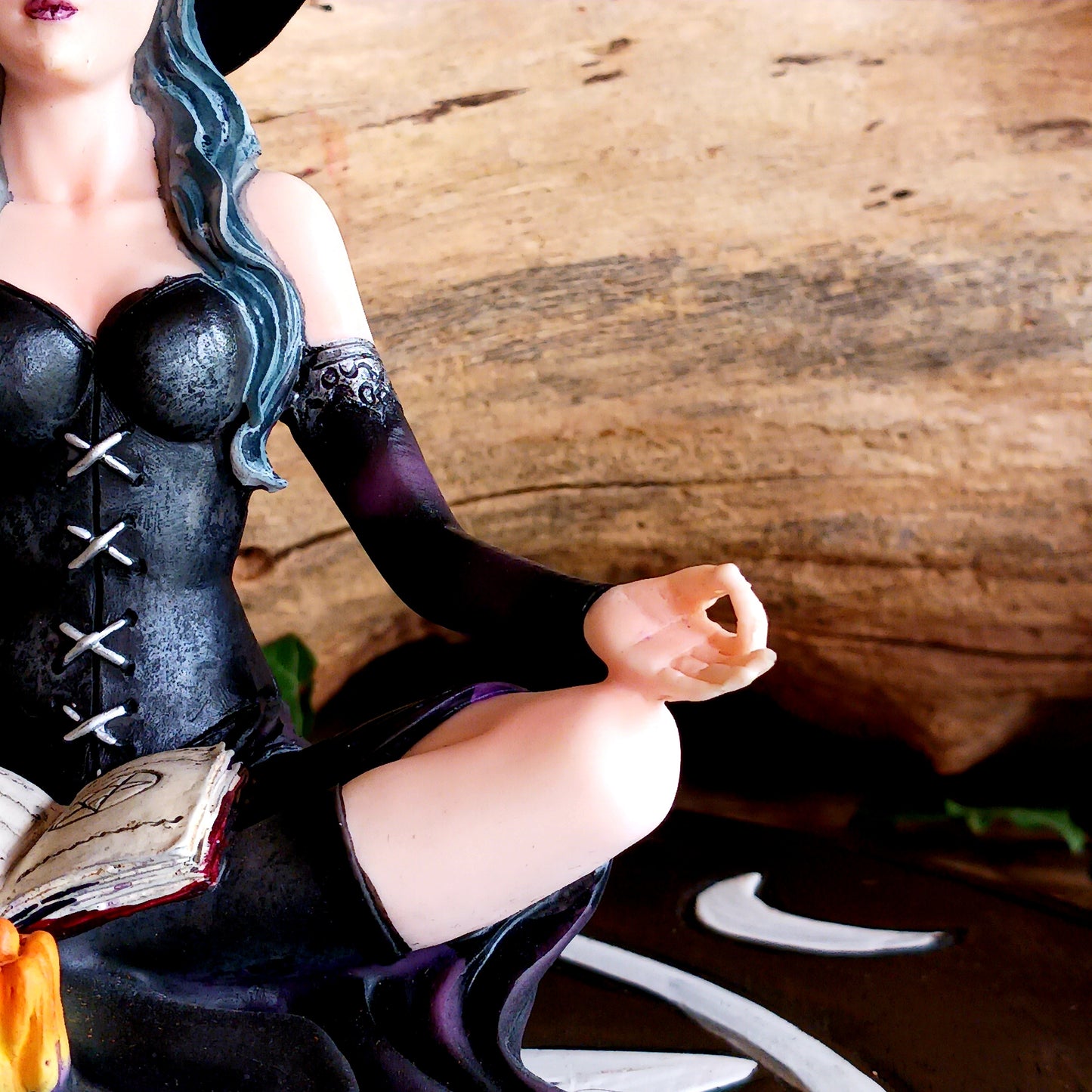 This stunning figurine features Aradia, wearing a black corseted dress, sitting crossed legged, cradling her grimoire with her legs, while staring into the burning flame of a candle.