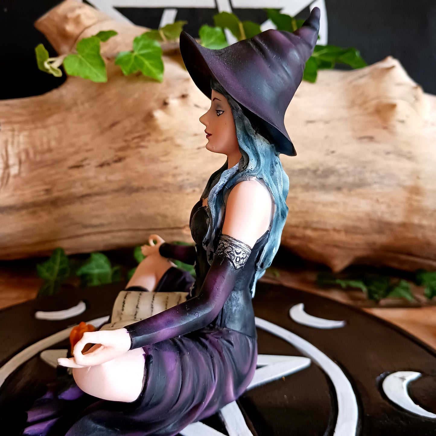 This stunning figurine features Aradia, wearing a black corseted dress, sitting crossed legged, cradling her grimoire with her legs, while staring into the burning flame of a candle.