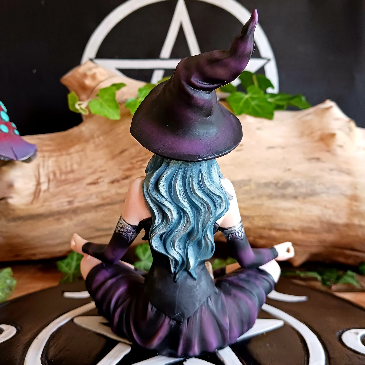 This stunning figurine features Aradia, wearing a black corseted dress, sitting crossed legged, cradling her grimoire with her legs, while staring into the burning flame of a candle.