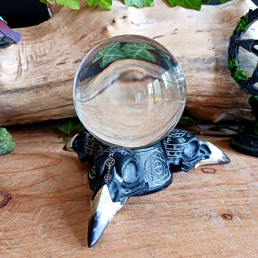 Crafted from high-quality resin and painted in a sophisticated black with exquisite gold detailing, this unique piece features three Raven skulls adorned with occult symbology. At the center of the crystal ball holder lies a pentacle surrounded by additional markings, creating an enchanting tableau. Resting atop the holder is a crystal ball, offering glimpses into unseen realms.