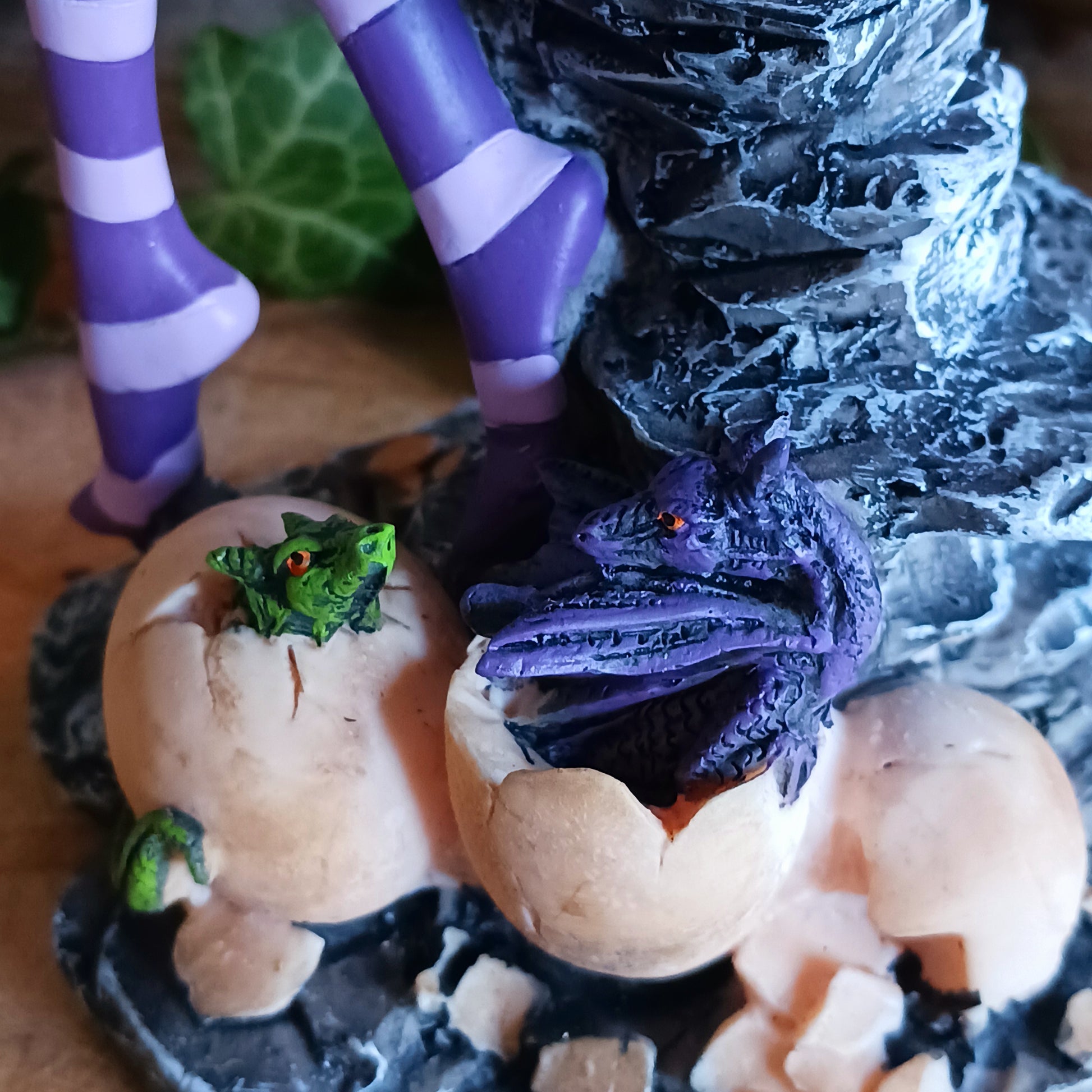 Sitting on a natural pillar of rock, this purple-winged Fairy looks to the side, blonde hair ending past her shoulders. On her shoulder, a purple Dragon hatchling looks around at the world cautiously, their green sibling curling defensively on the Fairy's lap. Around her feet, more Dragons hatch from bone-coloured eggs, taking their first look at the world. On the Fairy's forehead, a mystical symbol is marked in purple.