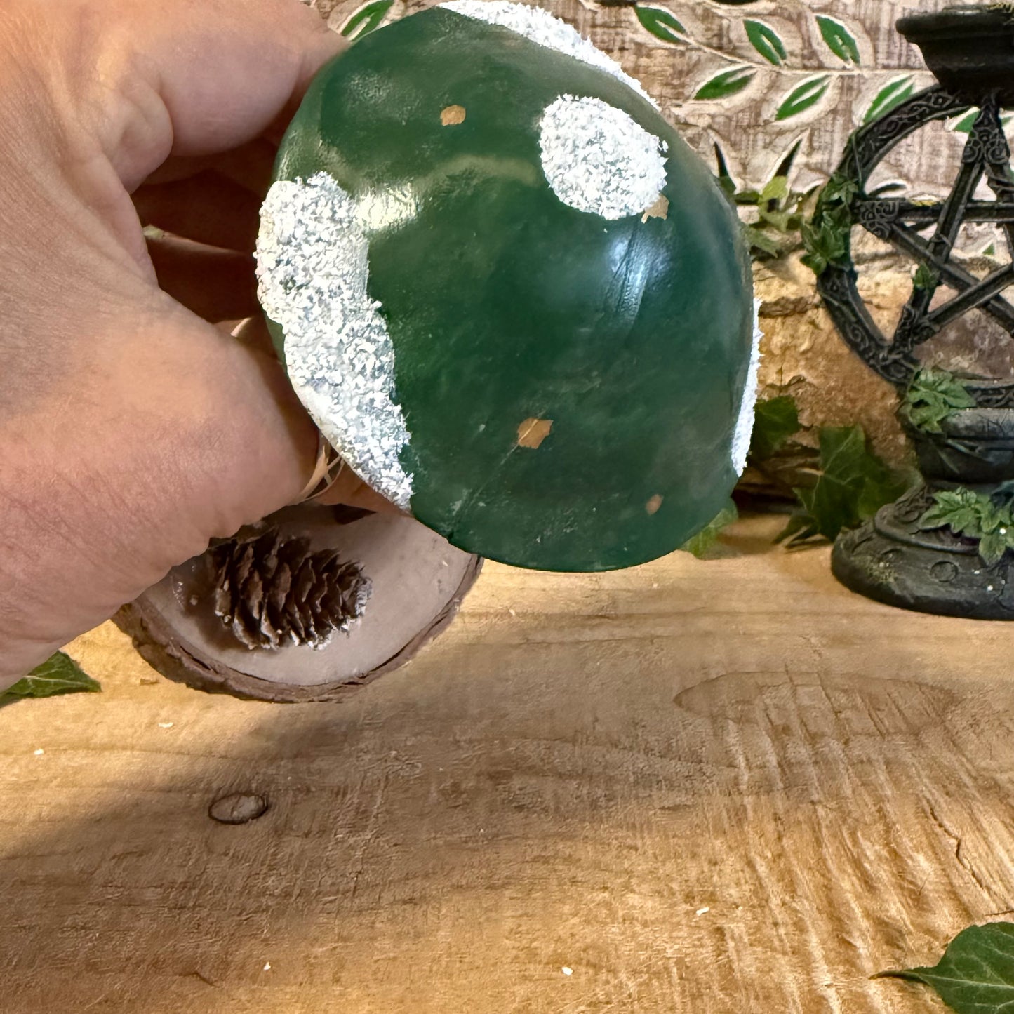 Hand holding a green and white decorative object on a wooden surface with plants in the background