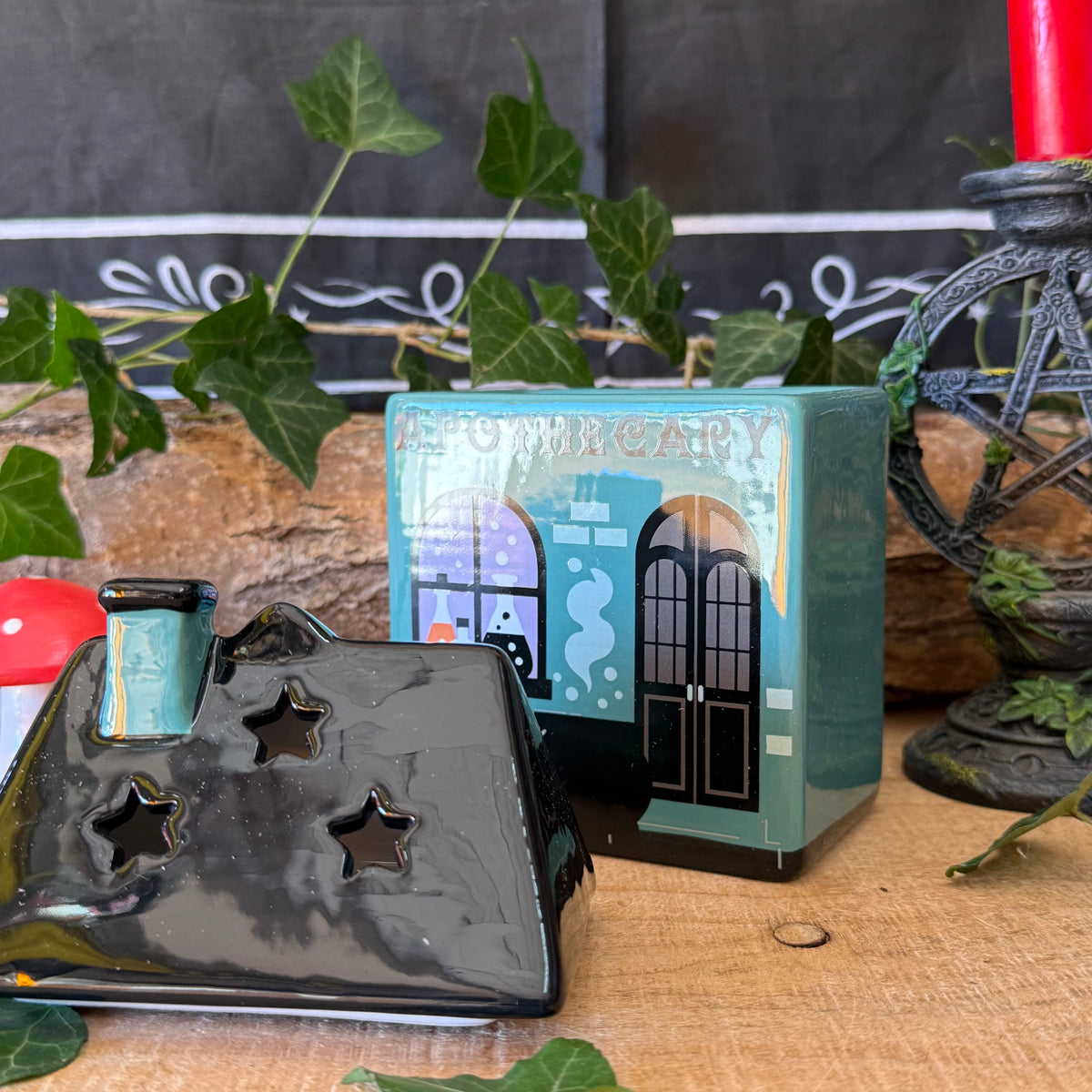 A decorative ceramic house-shaped candle holder with star-shaped cutouts sits in front of its matching illustrated turquoise-blue box on a wooden surface, with ivy and rustic decor in the background.