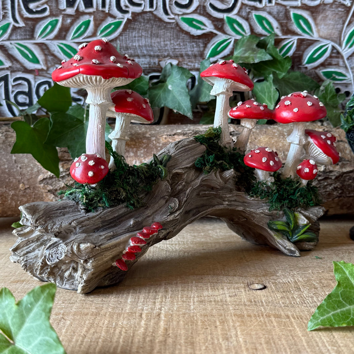 A decorative miniature mushroom cluster figurine featuring red and white gilled mushrooms growing on a weathered log base with faux moss and green ivy accents, displayed on a wooden surface.