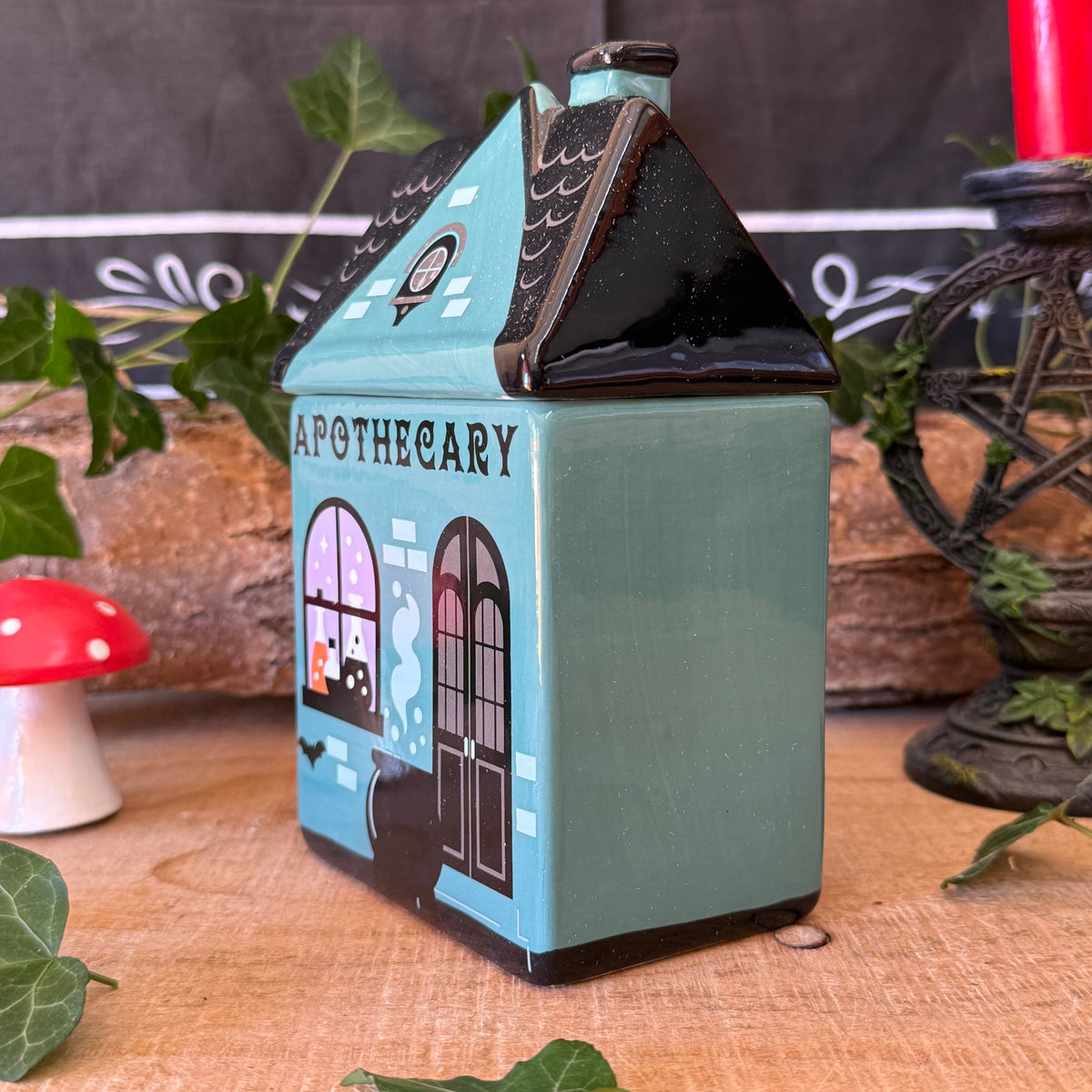 A small ceramic house-shaped apothecary jar with a teal body, black roof, and Halloween-themed window illustration, displayed on a wooden surface with decorative leaves, a mushroom, and a candle in the background.