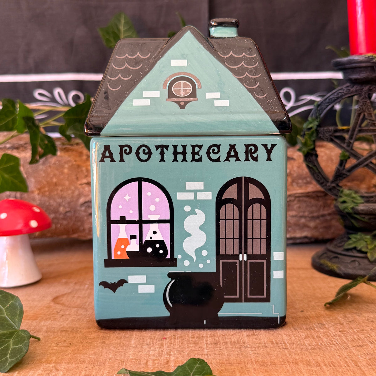 A small decorative house-shaped container labeled 'APOTHECARY' with a teal body, dark roof, illustrated window, cauldron, bats, and witch motifs, styled on a wooden surface with leaves and props around it.