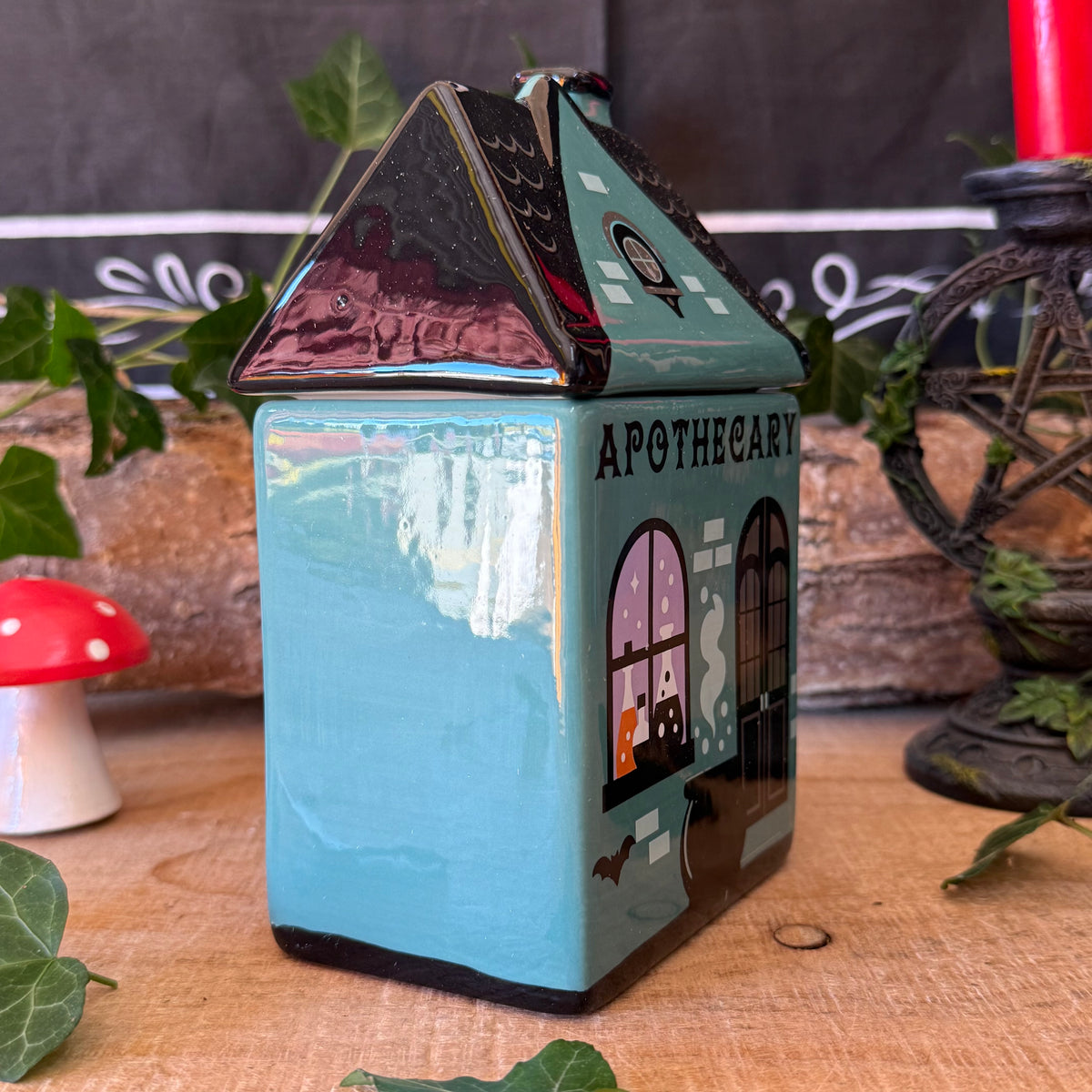 A small house-shaped ceramic jar or container with a turquoise body and dark roof, decorated with 'APOTHECARY' text and window illustrations. It is photographed on a wooden surface with decorative greenery, a mushroom, and a candle in the background.