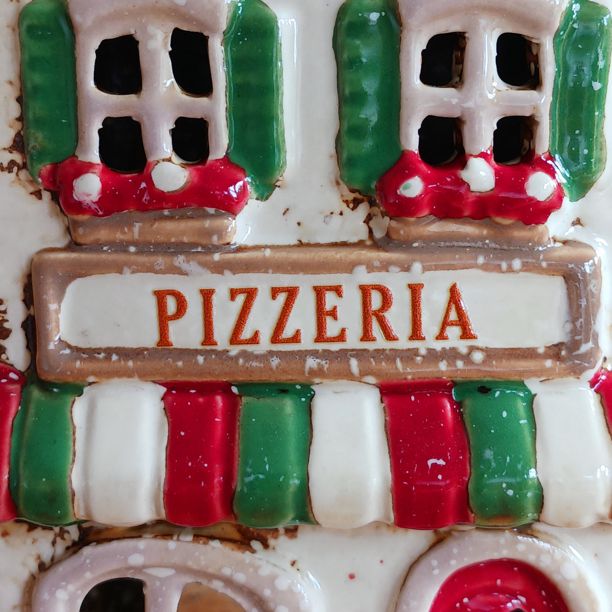 Bring a little taste of Italy into your home with this charming Village Pottery Pizzeria Tealight Holder. Designed as a delightful two-storey Italian village building, this cream pottery house features a brown tiled roof, chimney, green shutters, red flower boxes and colourful red and green striped awnings.