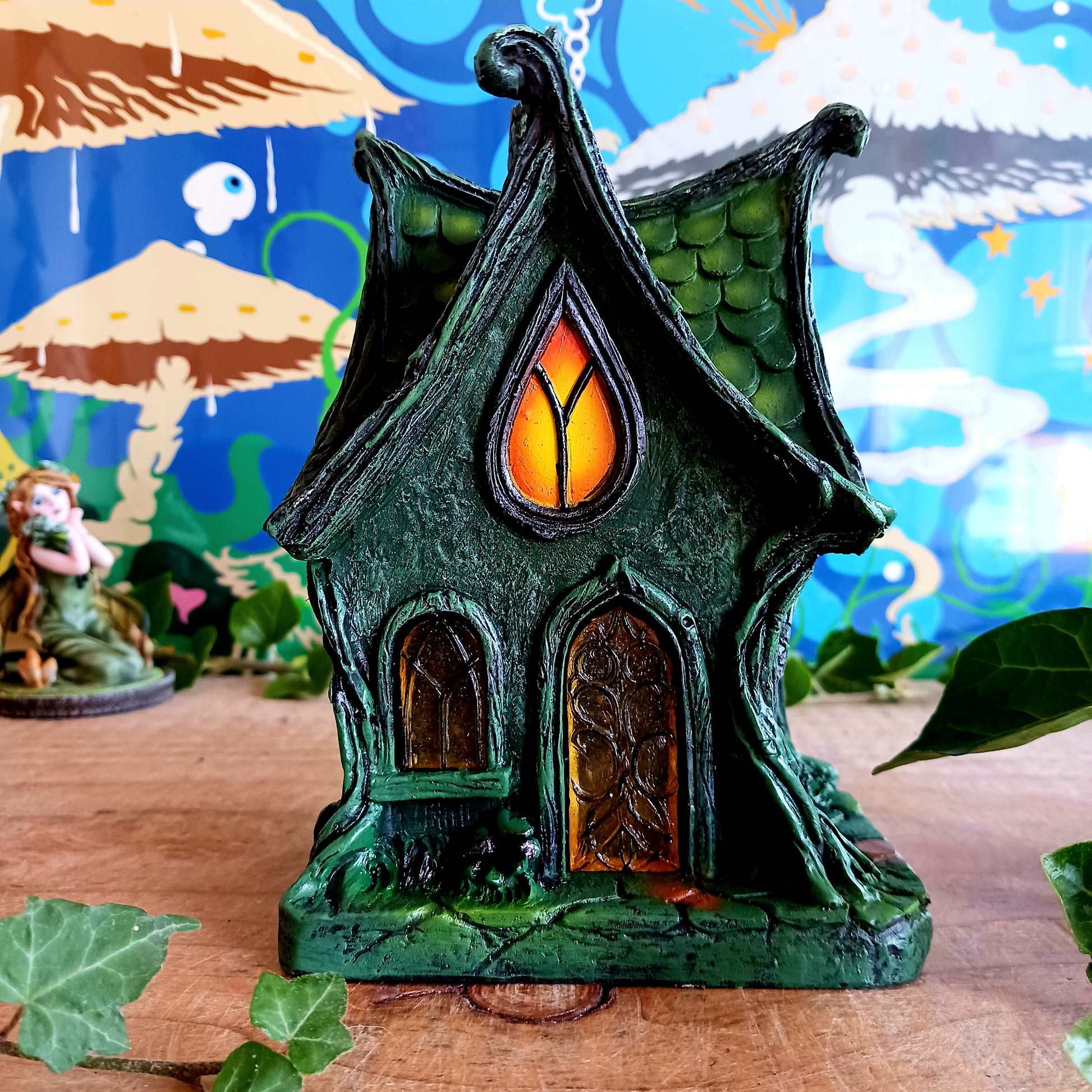 Step into a magical, enchanted forest with this beautiful gothic-style fairy cottage. Designed with a pointed roof adorned in dark green, scale-like shingles, this stunning piece glows warmly with LED lights shining through medieval-style windows. Sculpted vines and twisting tree roots climb the sides, while curled roof details add an intricate, whimsical finish.