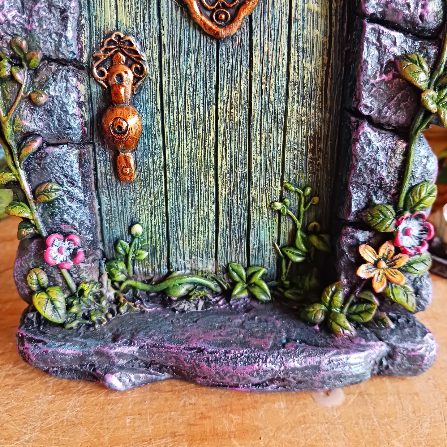 Step into a world of wonder with the Fae’s Forest Welcome fairy door. This beautifully crafted piece features a forest-green wooden-textured door framed by a detailed stone arch, adorned with twisting vines and bright red and yellow flowers.