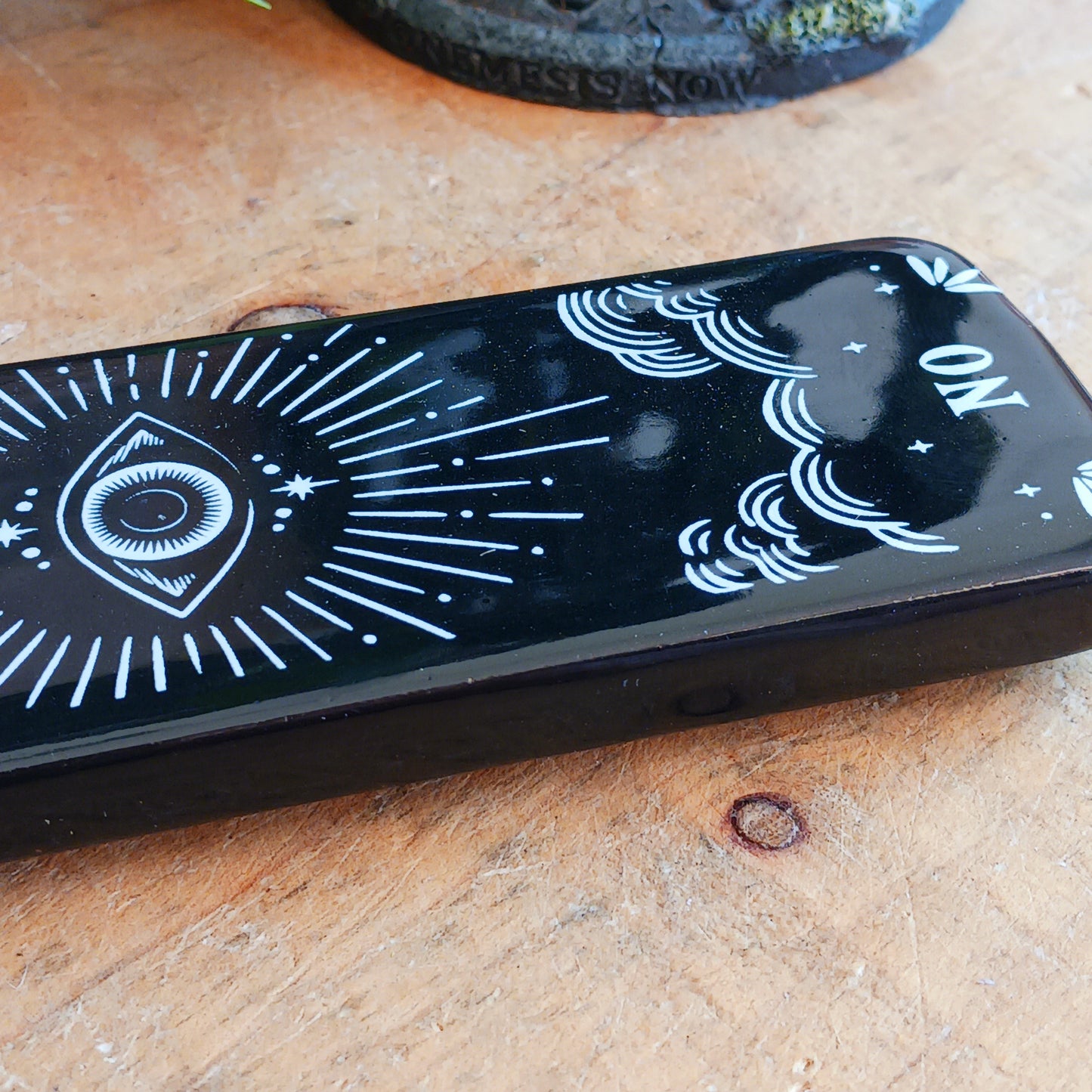 Add a touch of mystical gothic style to your incense rituals with this striking black incense ash catcher, inspired by traditional talking boards. Featuring a decorative planchette-style design surrounded by stars, moons and an all-seeing eye, it creates an atmospheric setting for burning your favourite incense sticks.