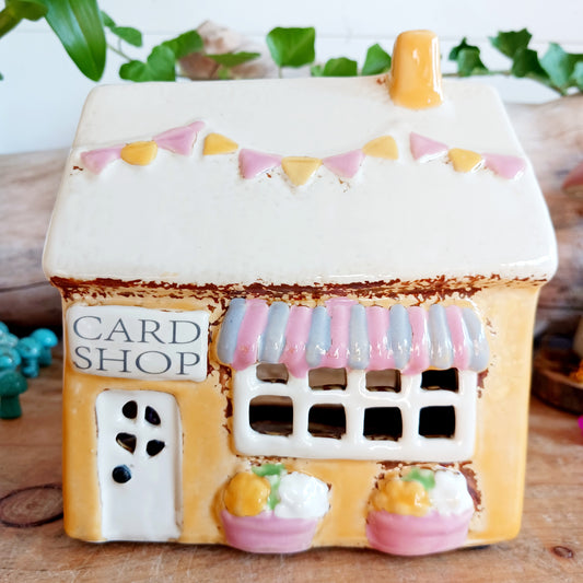 Add a whimsical touch to your décor with the Cards &amp; Gifts Tealight Holder from the Village Pottery Collection. Inspired by the charm of a cozy card shop in a quaint village, this ceramic piece features intricate detailing, from its bunting roof to its delicate windows. Perfect as a tealight holder or simply as a decorative ornament, it brings warmth and character to any space.