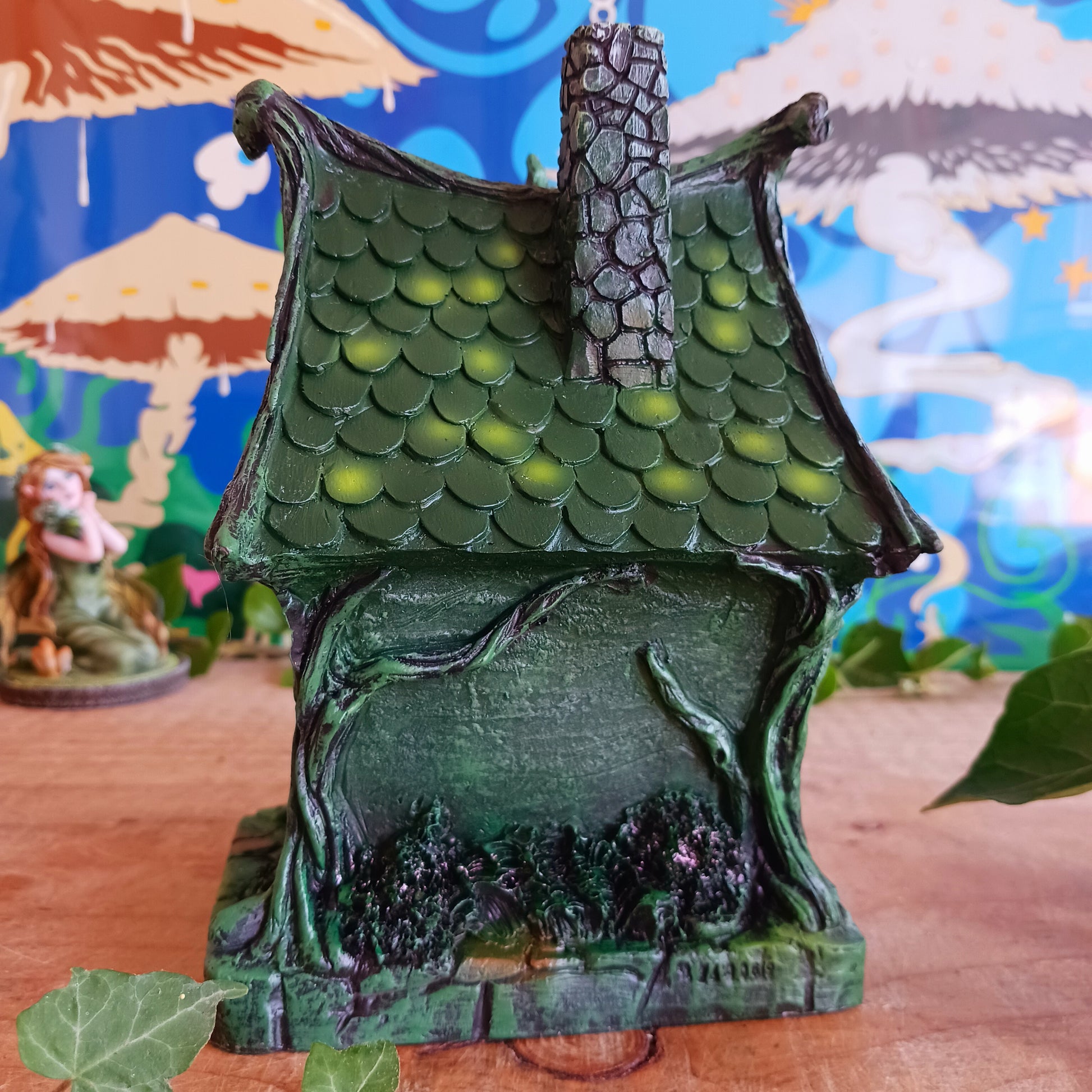 Step into a magical, enchanted forest with this beautiful gothic-style fairy cottage. Designed with a pointed roof adorned in dark green, scale-like shingles, this stunning piece glows warmly with LED lights shining through medieval-style windows. Sculpted vines and twisting tree roots climb the sides, while curled roof details add an intricate, whimsical finish.
