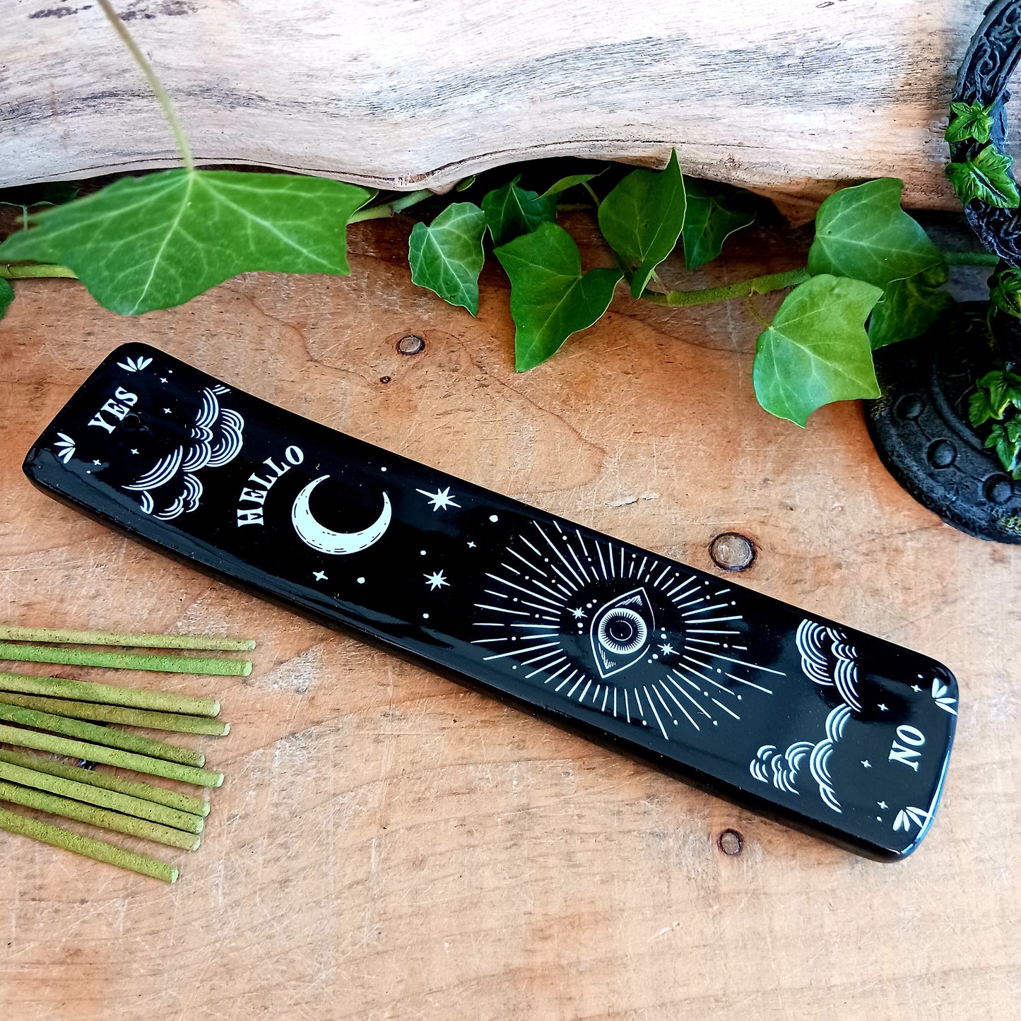 Add a touch of mystical gothic style to your incense rituals with this striking black incense ash catcher, inspired by traditional talking boards. Featuring a decorative planchette-style design surrounded by stars, moons and an all-seeing eye, it creates an atmospheric setting for burning your favourite incense sticks.