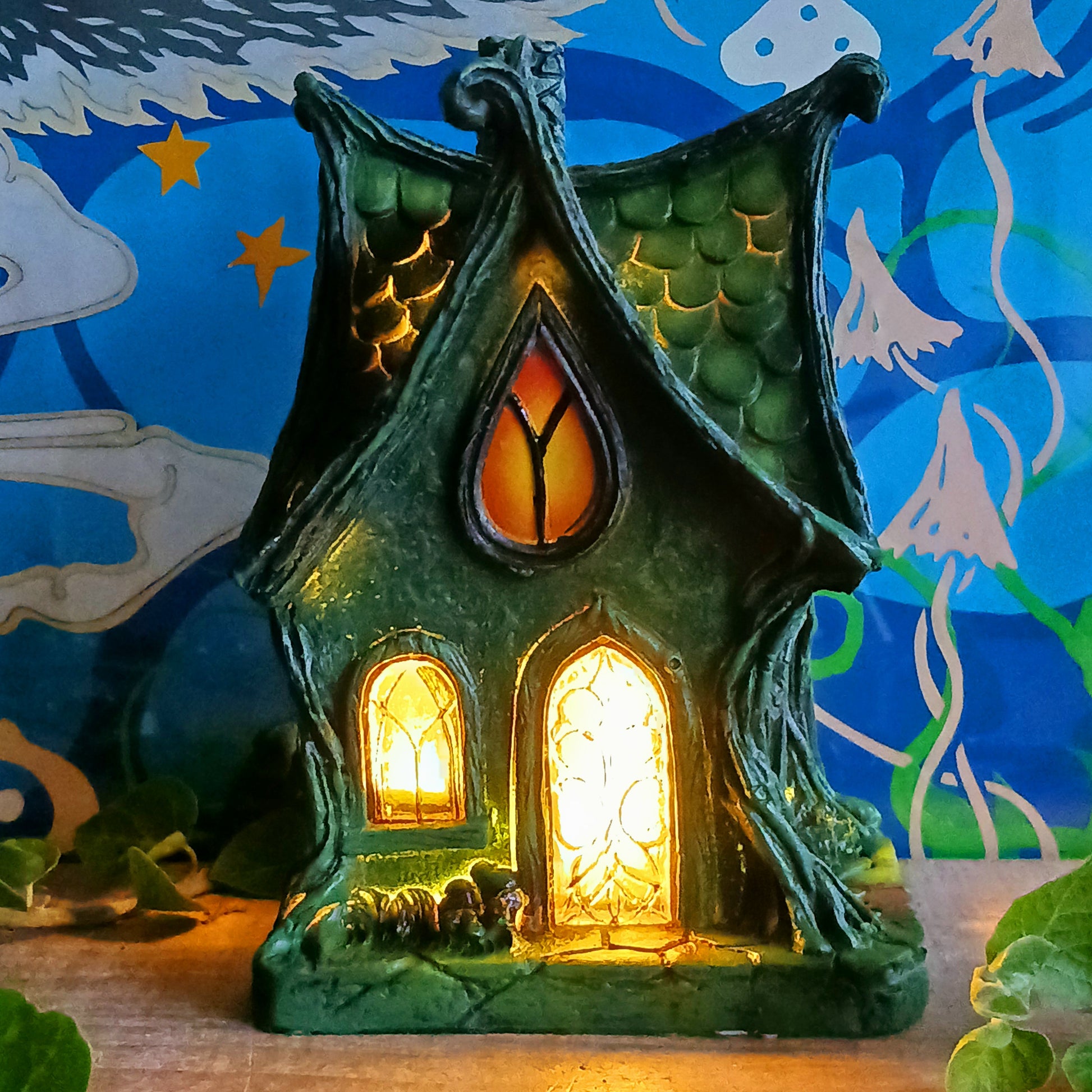 Step into a magical, enchanted forest with this beautiful gothic-style fairy cottage. Designed with a pointed roof adorned in dark green, scale-like shingles, this stunning piece glows warmly with LED lights shining through medieval-style windows. Sculpted vines and twisting tree roots climb the sides, while curled roof details add an intricate, whimsical finish.