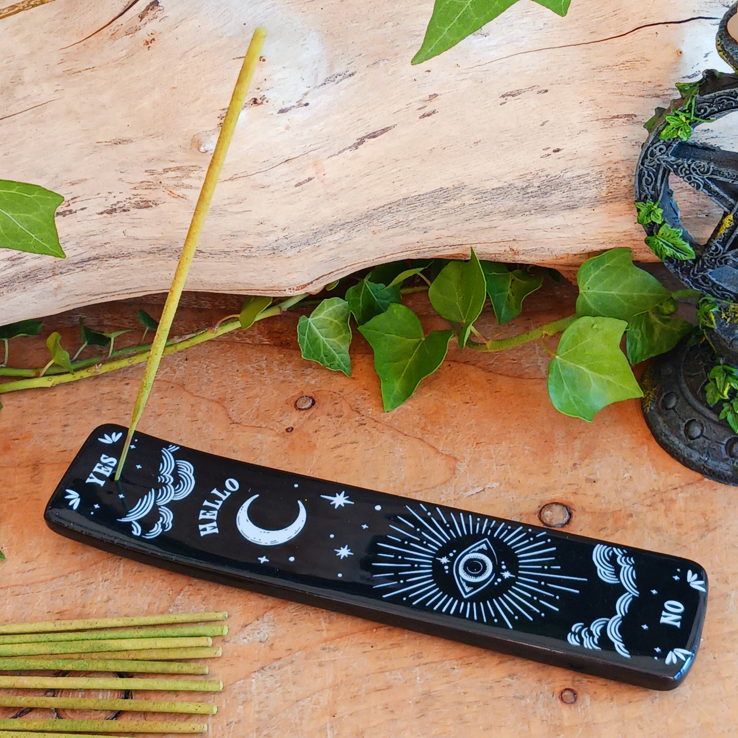 Add a touch of mystical gothic style to your incense rituals with this striking black incense ash catcher, inspired by traditional talking boards. Featuring a decorative planchette-style design surrounded by stars, moons and an all-seeing eye, it creates an atmospheric setting for burning your favourite incense sticks.