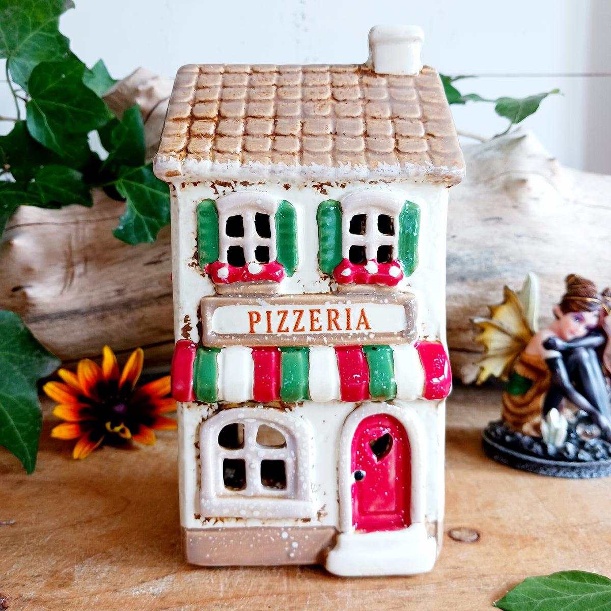 Bring a little taste of Italy into your home with this charming Village Pottery Pizzeria Tealight Holder. Designed as a delightful two-storey Italian village building, this cream pottery house features a brown tiled roof, chimney, green shutters, red flower boxes and colourful red and green striped awnings.