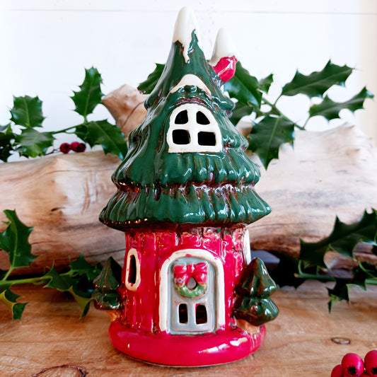 Step into a world of festive wonder with the Village Pottery Christmas Tree House Tealight Holder, a whimsical handcrafted ceramic creation glowing with the gentle light of the season. This enchanting piece blends the magic of a Christmas tree with the charm of a cosy cottage — complete with tiny windows, a detailed door, and a golden star crowning the roof.