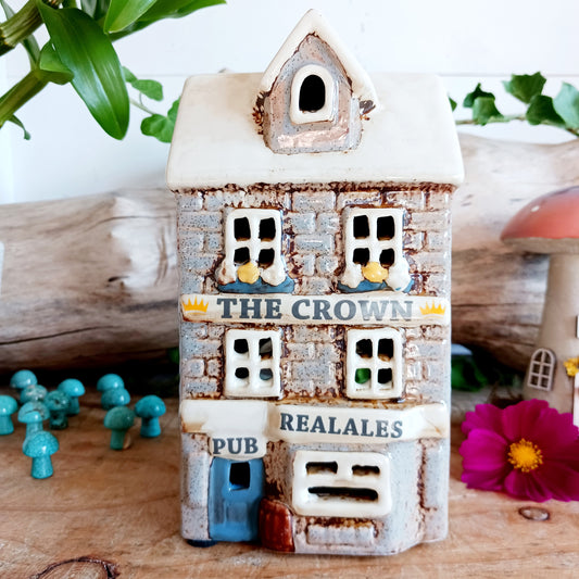 Bring the warmth of a cozy pub into your home with the Village Pottery Crown Pub Tealight. Crafted from ceramic with intricate detailing, this charming piece features a rear cut-out for easy placement of a tealight or string lights. Watch as the soft glow spills through the windows, creating a welcoming pub atmosphere. Perfect for building your own enchanting village display, this timeless decoration adds warmth and character to any space.