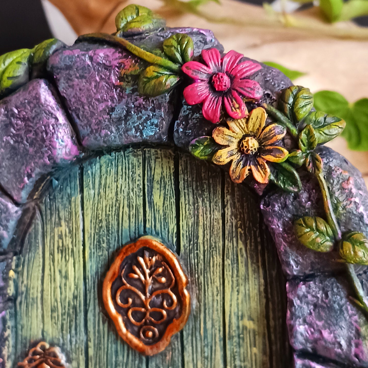 Step into a world of wonder with the Fae’s Forest Welcome fairy door. This beautifully crafted piece features a forest-green wooden-textured door framed by a detailed stone arch, adorned with twisting vines and bright red and yellow flowers.