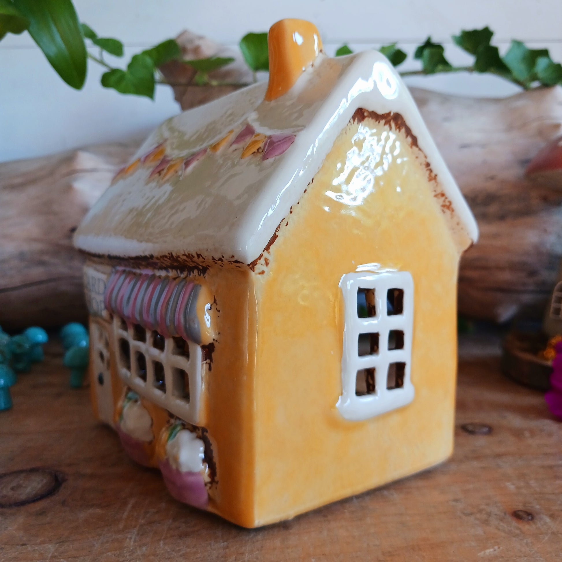 Add a whimsical touch to your décor with the Cards &amp; Gifts Tealight Holder from the Village Pottery Collection. Inspired by the charm of a cozy card shop in a quaint village, this ceramic piece features intricate detailing, from its bunting roof to its delicate windows. Perfect as a tealight holder or simply as a decorative ornament, it brings warmth and character to any space.