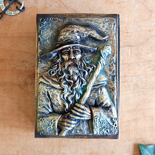 Bring a little magic and mystery into your home with the enchanting Wizard’s Hoard Box. Featuring a beautifully sculpted wizard clutching a magical staff, this decorative trinket box is surrounded by intricate mystical detailing and finished in a rich, antique gold-like colour.