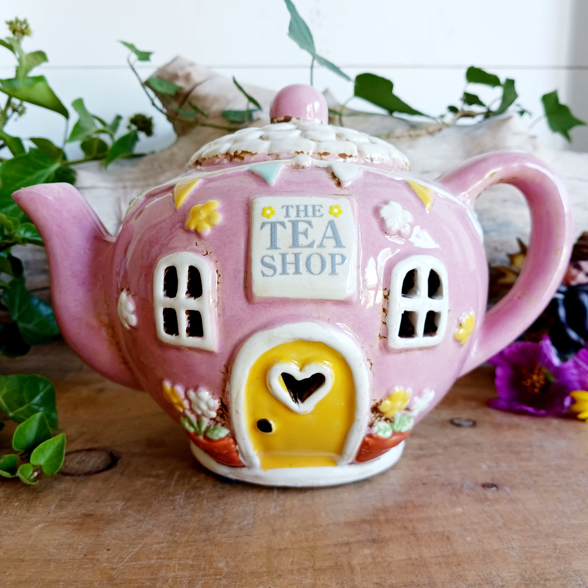 Add a sprinkle of charm to your home with this adorable Village Pottery ceramic tealight holder, beautifully designed as a whimsical pink teapot transformed into a cosy village tea shop. Featuring cottage-style windows, colourful bunting, pretty floral details and a welcoming heart-shaped doorway, this delightful decorative piece brings a touch of storybook magic to any room.