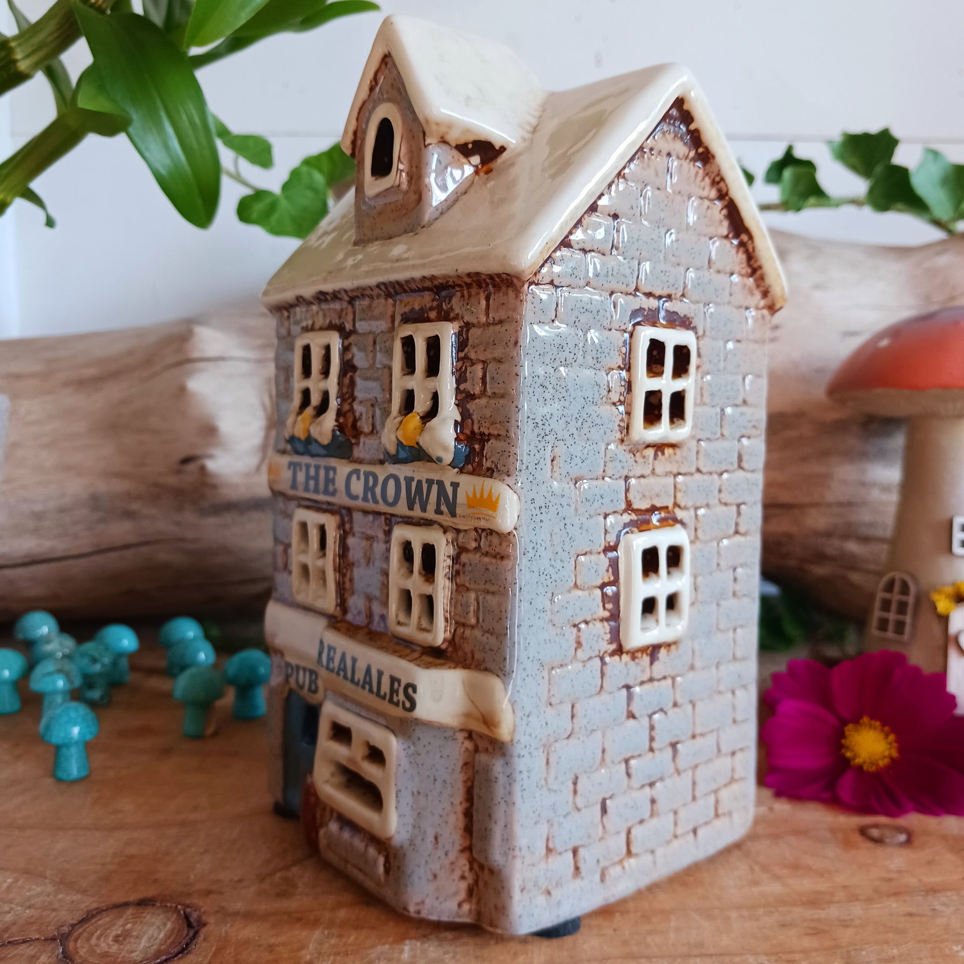 Bring the warmth of a cozy pub into your home with the Village Pottery Crown Pub Tealight. Crafted from ceramic with intricate detailing, this charming piece features a rear cut-out for easy placement of a tealight or string lights. Watch as the soft glow spills through the windows, creating a welcoming pub atmosphere. Perfect for building your own enchanting village display, this timeless decoration adds warmth and character to any space.