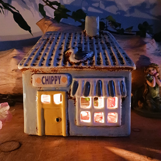 A warm, golden glow spills from the windows of this Village Pottery Chippy, where the spirit of a bustling village meets quiet enchantment. Crafted from ceramic, this Shudehill tealight house captures a familiar yet mystical charm, as candlelight flickers like stories shared after dusk.