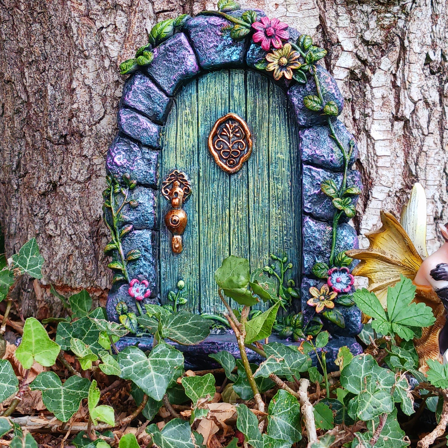 Step into a world of wonder with the Fae’s Forest Welcome fairy door. This beautifully crafted piece features a forest-green wooden-textured door framed by a detailed stone arch, adorned with twisting vines and bright red and yellow flowers.