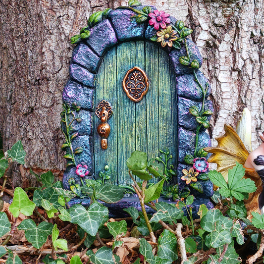 Step into a world of wonder with the Fae’s Forest Welcome fairy door. This beautifully crafted piece features a forest-green wooden-textured door framed by a detailed stone arch, adorned with twisting vines and bright red and yellow flowers.