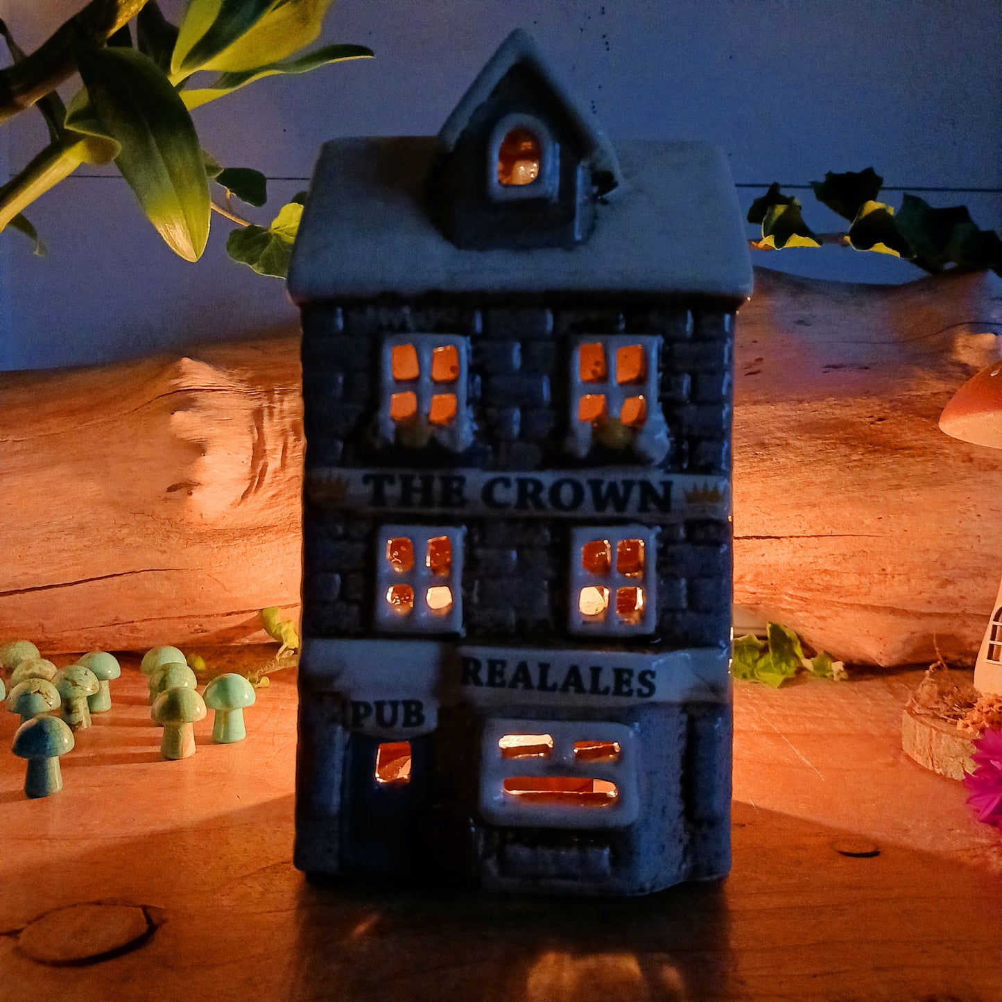 Bring the warmth of a cozy pub into your home with the Village Pottery Crown Pub Tealight. Crafted from ceramic with intricate detailing, this charming piece features a rear cut-out for easy placement of a tealight or string lights. Watch as the soft glow spills through the windows, creating a welcoming pub atmosphere. Perfect for building your own enchanting village display, this timeless decoration adds warmth and character to any space.