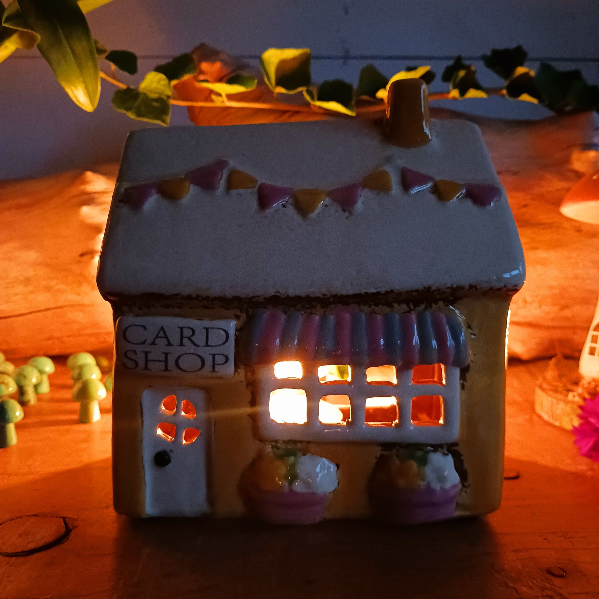 Add a whimsical touch to your décor with the Cards &amp; Gifts Tealight Holder from the Village Pottery Collection. Inspired by the charm of a cozy card shop in a quaint village, this ceramic piece features intricate detailing, from its bunting roof to its delicate windows. Perfect as a tealight holder or simply as a decorative ornament, it brings warmth and character to any space.