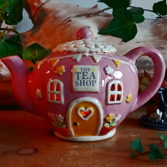 Add a sprinkle of charm to your home with this adorable Village Pottery ceramic tealight holder, beautifully designed as a whimsical pink teapot transformed into a cosy village tea shop. Featuring cottage-style windows, colourful bunting, pretty floral details and a welcoming heart-shaped doorway, this delightful decorative piece brings a touch of storybook magic to any room.