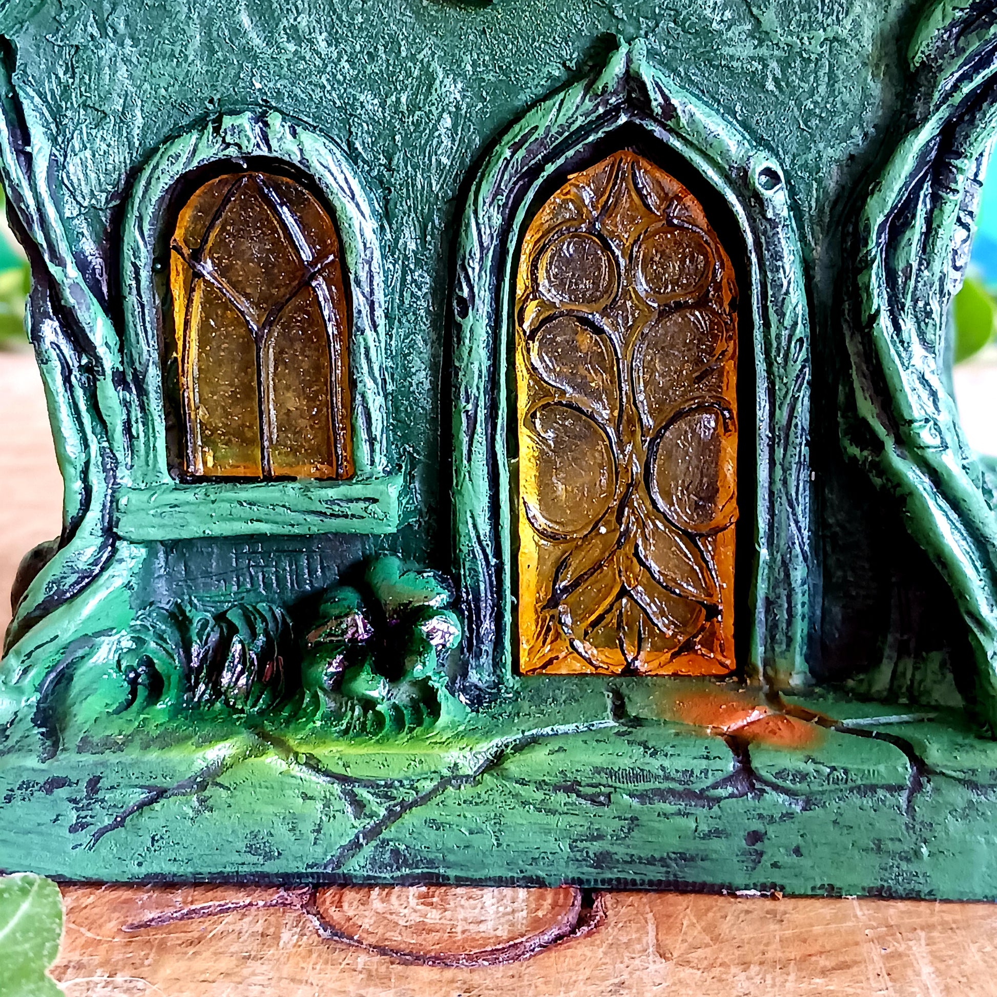 Step into a magical, enchanted forest with this beautiful gothic-style fairy cottage. Designed with a pointed roof adorned in dark green, scale-like shingles, this stunning piece glows warmly with LED lights shining through medieval-style windows. Sculpted vines and twisting tree roots climb the sides, while curled roof details add an intricate, whimsical finish.