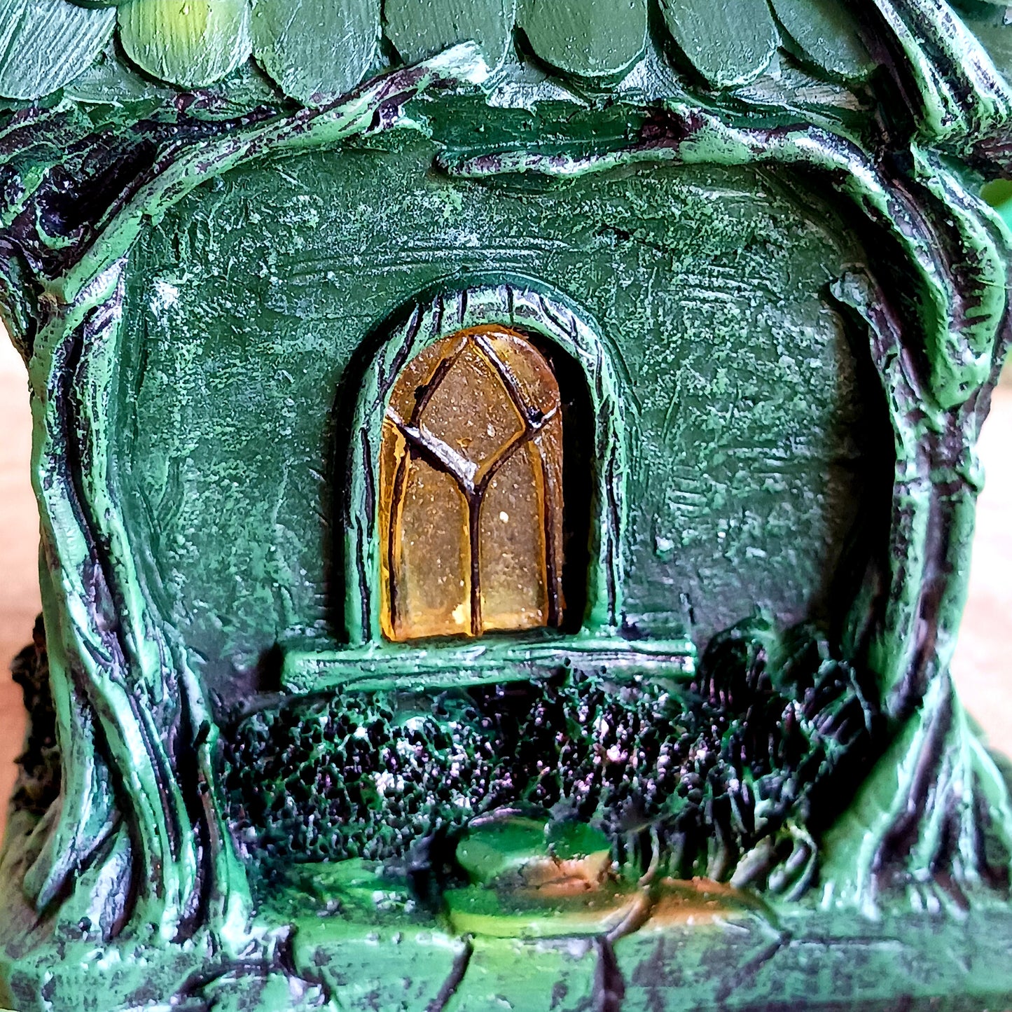 Step into a magical, enchanted forest with this beautiful gothic-style fairy cottage. Designed with a pointed roof adorned in dark green, scale-like shingles, this stunning piece glows warmly with LED lights shining through medieval-style windows. Sculpted vines and twisting tree roots climb the sides, while curled roof details add an intricate, whimsical finish.